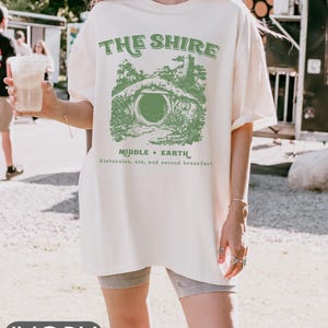 Pode incluir: Uma camiseta branca oversized com um gr&aacute;fico verde de uma toca de hobbit e o texto "The Shire, Middle-Earth, Elevensies, ale, and second breakfast".
