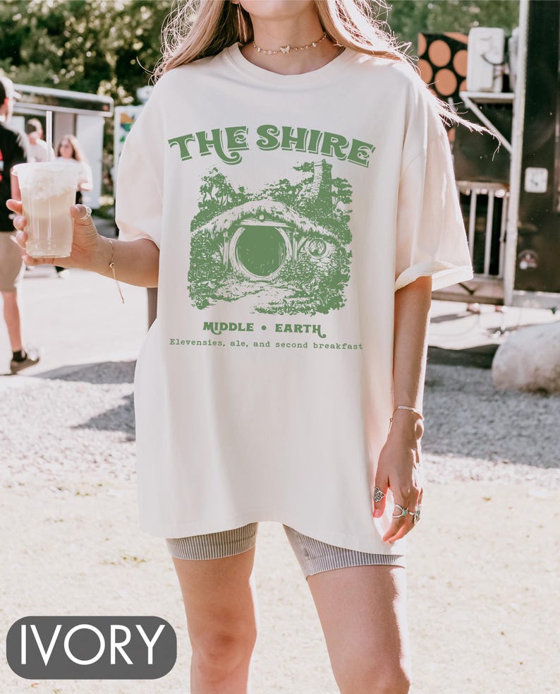 Pode incluir: Uma camiseta branca oversized com um gr&aacute;fico verde de uma toca de hobbit e o texto "The Shire, Middle-Earth, Elevensies, ale, and second breakfast".