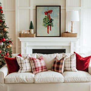 May include: A cozy living room scene with a white sofa adorned with red and plaid pillows. A framed mistletoe print hangs above a fireplace. A Christmas tree with red and gold ornaments is on the left.