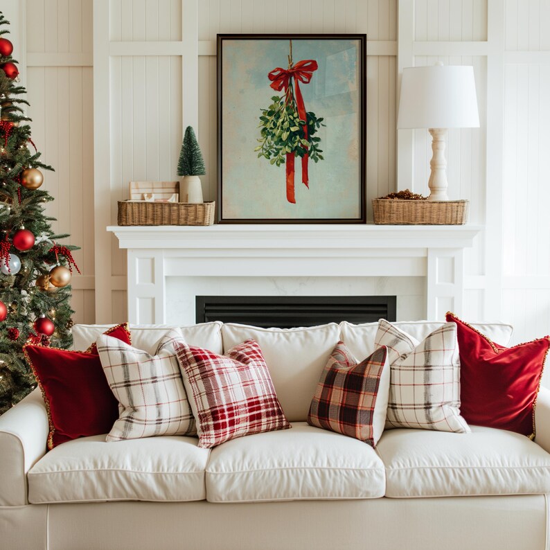 May include: A cozy living room scene with a white sofa adorned with red and plaid pillows. A framed mistletoe print hangs above a fireplace. A Christmas tree with red and gold ornaments is on the left.