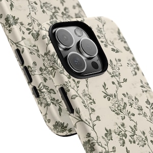 Puede incluir: Primer plano de dos fundas de tel&eacute;fono con un estampado floral. Las fundas son de color blanco roto con un aspecto envejecido y presentan un dise&ntilde;o repetido de flores y enredaderas verdes. La funda del tel&eacute;fono tiene un protector de lente negro.