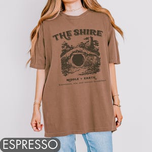 Pode incluir: Uma camiseta marrom oversized com uma estampa gr&aacute;fica de uma toca de hobbit e o texto "The Shire, Middle-Earth, Elevensies, ale, and second breakfast".