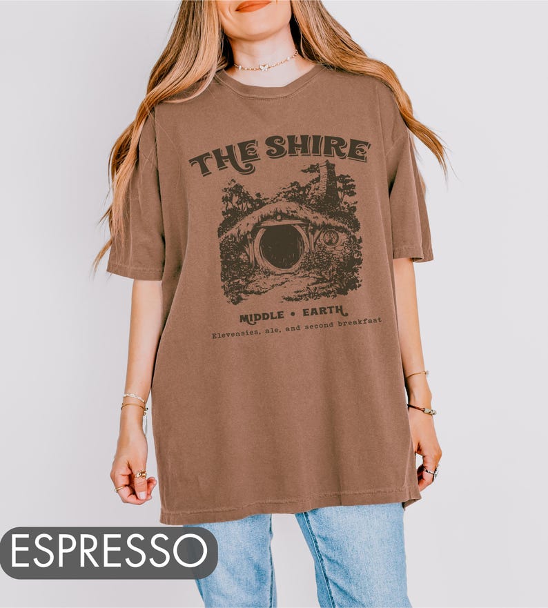 Pode incluir: Uma camiseta marrom oversized com uma estampa gr&aacute;fica de uma toca de hobbit e o texto "The Shire, Middle-Earth, Elevensies, ale, and second breakfast".