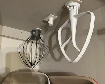 Op de afbeelding: Witte keukenmixer-accessoires die aan haken aan een wit kastje hangen. De accessoires omvatten een garde en een deeghaak. Een witte keramische bakvorm staat op de voorgrond.