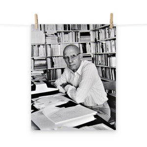 May include: A black and white photo of a man wearing glasses sitting at a desk in a library. He is surrounded by bookshelves filled with books. He is looking at the camera.