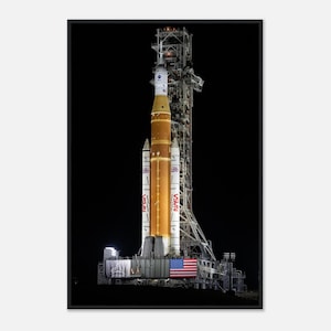 May include: A vertical image of the NASA Space Launch System (SLS) rocket on its launchpad, illuminated against a black sky. The rocket is primarily orange and white, with the American flag visible. The launch tower is also visible.