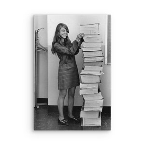 May include: A monochrome photograph of a woman in a striped dress and glasses, smiling as she organises a high pile of books. The image, likely from the 1960s or 1970s, suggests a library or academic environment, with a vintage aesthetic.