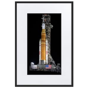May include: A framed print featuring a NASA rocket on a launch pad at night. The rocket is primarily orange and white, with the American flag visible. The launch tower is visible in the background against a black sky.
