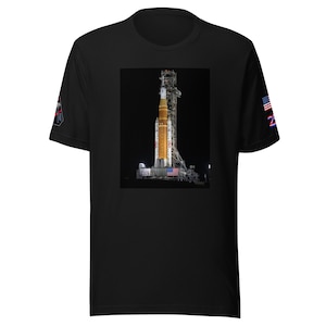 May include: Black t-shirt featuring a vibrant image of a rocket on a launchpad at night, with an American flag. The rocket is orange and white. The shirt has a small patch on the left sleeve and a design on the right sleeve.