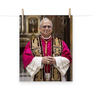 Puede incluir: Impresión enmarcada de un hombre con vestimenta religiosa. Viste una túnica magenta con bordados dorados y un cordón dorado. Las mangas son blancas con detalles intrincados. Sonríe y lleva gafas.