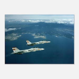 May include: Two light gray fighter jets flying in formation over a deep blue body of water. The aircraft feature swept-back wings, dual vertical stabilizers, and red and white markings on the tail sections, set against a coastal landscape.