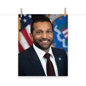 Könnte beinhalten: Ein Porträt eines Mannes in einem dunkelblauen Anzug mit roter Krawatte. Er lächelt und hat einen Bart. Der Hintergrund ist ein verschwommenes Bild der amerikanischen Flagge und eines Siegels.