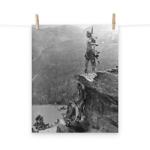 May include: Black and white photograph of three Native American people standing on a rocky cliff overlooking a valley. The person in the center is standing tall and holding a spear, while the two people on the left are sitting down. The photo is taken from a low angle, giving the viewer a sense of the vastness of the landscape.