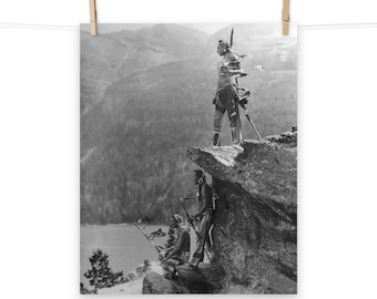 May include: Black and white photograph of three Native American people standing on a rocky cliff overlooking a valley. The person in the center is standing tall and holding a spear, while the two people on the left are sitting down. The photo is taken from a low angle, giving the viewer a sense of the vastness of the landscape.