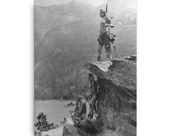 May include: Black and white photograph of three Native American people standing on a rocky cliff overlooking a lake and mountains. The people are wearing traditional clothing and holding weapons.