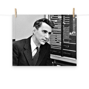 May include: A black and white photo of a man in a suit and tie, looking to the left. He is standing in front of a large computer panel with lights.