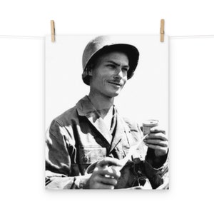 May include: A black and white photo of a man in a military uniform holding a can. He is wearing a helmet and has a serious expression on his face.