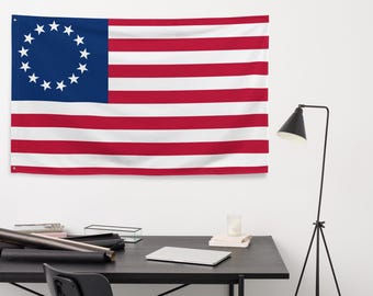 Pode incluir: Uma bandeira americana vermelha, branca e azul com 13 listras e um círculo de 13 estrelas no cantão. A bandeira está pendurada em uma parede branca acima de uma mesa preta com um laptop, um porta-velas de vidro e uma lâmpada preta.