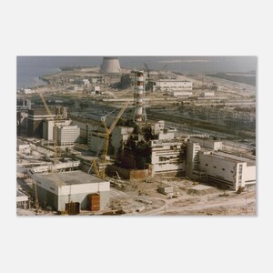 Op de afbeelding: Een luchtfoto van een kerncentralecomplex in aanbouw. De afbeelding toont meerdere gebouwen, een hoge schoorsteen met rood-witte strepen en een koeltoren. Bouwkranen zijn zichtbaar op de bouwplaats.