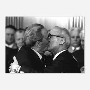 May include: Black and white photograph of two men embracing and kissing. Both men are wearing suits and ties. Other men are visible in the background, out of focus.