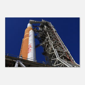 May include: A large rocket, predominantly orange and white, bearing the NASA logo, is positioned next to a complex launch tower. The structure is set against a deep blue sky. The launch tower is constructed from metal and features numerous levels.
