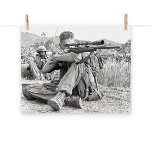 May include: A black and white photograph of a soldier aiming a rifle with a scope. The soldier is sitting on the ground with a backpack on. Another soldier is sitting behind him.