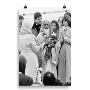 Pode incluir: Uma fotografia a preto e branco de um homem com uma longa barba branca e cabelo branco, vestindo uma túnica branca e segurando um ramo de flores. Ele está rodeado de pessoas, algumas das quais estão vestindo casacos.