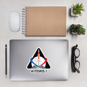 Może przedstawiać: Naklejka z logo misji Artemis I. Projekt przedstawia rakietę startującą z księżyca, z niebieskimi i czerwonymi ścieżkami orbitalnymi. Tekst "ARTEMIS I" jest wyświetlany poniżej. Naklejka znajduje się na laptopie.