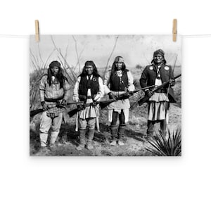 Op de afbeelding: Vier inheemse Amerikaanse mannen staan op een veld, elk met een geweer in de hand. Ze dragen traditionele kleding en hoofddeksels. De foto is in zwart-wit.