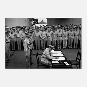 May include: Black and white photograph of a historical event. A seated officer signs documents at a table, surrounded by uniformed personnel. A ship is visible in the background, with a large group of officers standing behind the table.