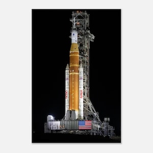 May include: A tall, multi-stage rocket, the Space Launch System (SLS), is positioned on a launch pad. The rocket is primarily orange and white, with the NASA logo visible. An American flag is displayed near the base of the rocket, set against a black background.