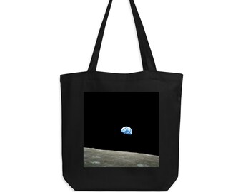 Op de afbeelding: Een zwarte tote bag met een witte afbeelding van de aarde die opkomt boven het oppervlak van de maan.