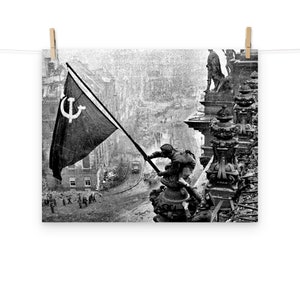 Puede incluir: Una fotografía en blanco y negro de un soldado izando una bandera roja con un martillo y una hoz blancos en un edificio en una ciudad devastada por la guerra. El soldado está de pie en un edificio dañado con otros soldados en el fondo.