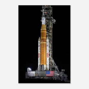 May include: A tall, orange and white NASA rocket, the Space Launch System (SLS), stands on a launch pad. The rocket is flanked by two white boosters and the American flag is visible. The background is a dark night sky.