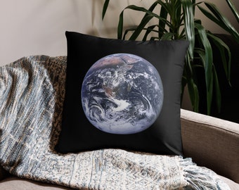 May include: A black throw pillow with a blue and white image of Earth on it. The pillow is sitting on a couch with a blue and white throw blanket.