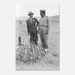 May include: A monochrome photograph showing two men in a desolate environment. One is in a suit and hat, the other in a military uniform. A metal rod structure is in the foreground.