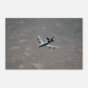 May include: A gray military surveillance aircraft with four engines and a large, circular radar dome mounted on top of the fuselage, flying over a vast, textured, and arid landscape with scattered patches of light-colored terrain.