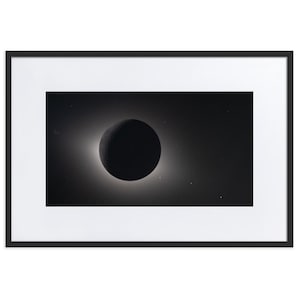May include: A framed black and white print of a solar eclipse. The image features a dark, circular shape in the center, surrounded by a bright halo. The print is set in a black frame with a white mat.