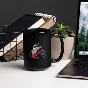May include: A black ceramic mug featuring a triangular design with a stylized moon and planet graphic, red and white accents, and text. The mug is on a white surface with a laptop, books, and a plant in the background.