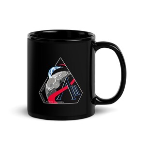 May include: Black ceramic mug with a space-themed design. The design features a stylized moon, Earth, and a geometric shape with the names WISEMAN, GLOVER, KOCH, and HANSEN. The mug has a glossy finish and a curved handle.