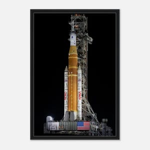 May include: A framed photograph of the NASA Artemis I rocket on its launchpad. The rocket is orange and white, with the NASA logo visible. The American flag is displayed at the base of the rocket.
