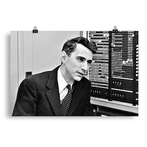 Op de afbeelding: Een zwart-witfoto van een man in pak en stropdas, die naar rechts kijkt. Hij staat voor een groot computerpaneel.