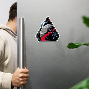 May include: A triangular sticker featuring a space-themed design. The design includes a stylized planet, a crescent moon, and the letter "A" in red. The names "Wiseman", "Glover", "Koch", and "Hansen" are printed around the edges.