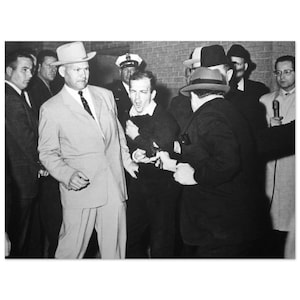 Może przedstawiać: Czarno-białe zdjęcie grupy mężczyzn, z których niektórzy są w garniturach i kapeluszach, otaczających mężczyznę w czarnym swetrze. Mężczyzna w swetrze wydaje się krzyczeć, podczas gdy inni go trzymają. Mężczyzna w jasnym garniturze i kapeluszu jest na pierwszym planie.