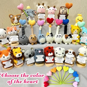 May include: A collection of handmade crochet animal figurines, including cats, cows, and sloths, each holding a heart-shaped accessory. The figurines are displayed on wooden bases, with small signs. The text "Choose the color of the heart" is at the bottom.