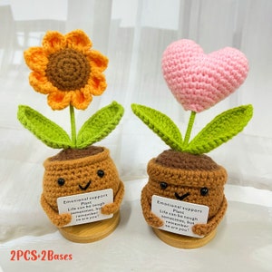 May include: Two crocheted plant pots with a sunflower and a heart on top. Each pot has a smiling face and is holding a card that reads "Emotional support plant. Life can be tough sometimes, but remember so are you!"