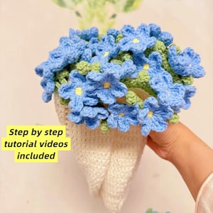 May include: A handmade crochet flower arrangement with light blue flowers, yellow centres, and green leaves, in a white crochet vase. The image includes the text "Step by step tutorial videos included."