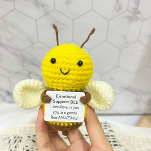 Op de afbeelding: Een handgemaakte gehaakte bij met een gele body, bruine accenten en crèmekleurige vleugels. De bij houdt een bordje vast met de tekst "Emotional Support BEE" en een bemoedigende boodschap. Een schattig, troostrijk item.