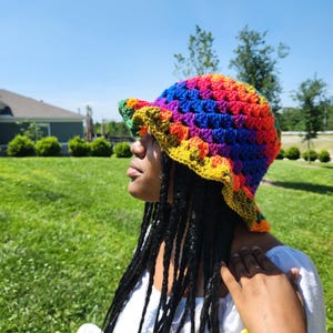 May include: A person wearing a rainbow crochet bucket hat with a white shirt and colorful bracelets. The hat has a wide brim and is made with a variety of colors, including red, orange, yellow, green, blue, and purple.
