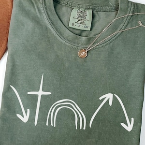 以下が含まれることがあります： オリーブグリーンのComfort Colors Tシャツ。白い十字架、虹、矢印のグラフィックが描かれています。小さなペンダントが付いたゴールドカラーのネックレスがシャツの上にあります。シャツのサイズはSです。